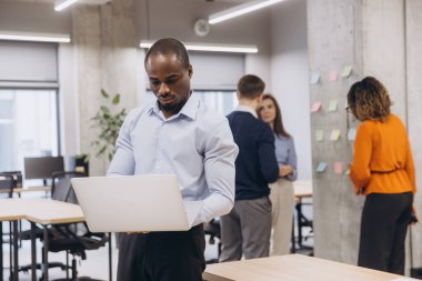 Modern ofis ortamında birlikte çalışan, tartışmalara katılan ve teknolojiyi üretkenliği arttırmak için kullanan, çeşitli profesyonel dizüstü bilgisayar kullanan Afrikalı Amerikalı bir adam.