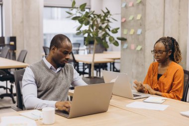 Profesyonel Afro-Amerikalı meslektaşlar iş birliği yapıyor, dizüstü bilgisayarlarda daktilo yazıyor ve çağdaş çalışma alanı içindeki evrakları inceliyor