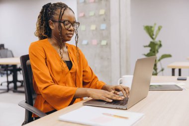 Profesyonel Afro-Amerikalı iş kadını notlar ve bitkilerle çevrili modern ofis ortamında özenle bir dizüstü bilgisayar üzerinde çalışıyor.