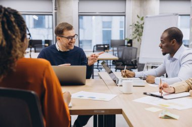 Dizüstü bilgisayarlar ve çizelgeler kullanarak, çağdaş bir çalışma alanında takım çalışması ve çeşitliliğin gösterildiği, işbirlikçi bir toplantıya katılan profesyoneller