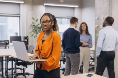 Laptop kullanan Afrikalı Amerikalı bir kadın, çağdaş bir ofiste birlikte çalışan profesyoneller, çeşitlilik ve takım çalışması sergiliyorlar. Teknoloji ve işbirliğine odaklan.