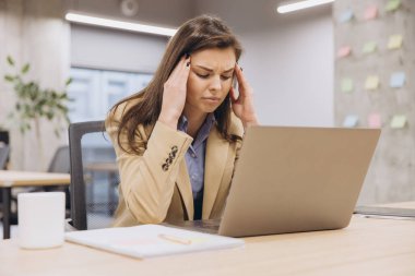 Modern ofis ortamında dizüstü bilgisayarı üzerinde çalışırken baş ağrısı çeken genç iş kadını, teslim tarihlerinin baskısını hissediyor.