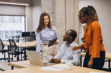 Profesyonel ekip, modern, iyi aydınlatılmış ofis ortamında dizüstü bilgisayar ve dijital cihazlar kullanırken proje detaylarını tartışıyor.