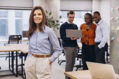 Profesyonel iş kadını güvenle ayakta duruyor, elleri cebinde, çağdaş kurumsal çalışma alanında işbirliği içinde çalışan bir ekiple birlikte.