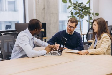 Profesyoneller çağdaş bir ofiste ortak bir toplantıda dizüstü bilgisayarlar ve belgelerle stratejileri tartışıyor, takım çalışmasını ve yenilikleri teşvik ediyor