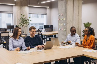 Dört meslektaş birlikte çalışarak dizüstü bilgisayarlar ve tabletler kullanıyor, fikirleri paylaşıyor ve modern ofis alanlarında stratejileri tartışıyorlar.