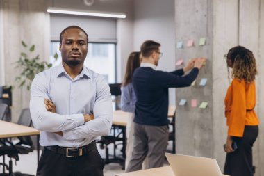 Çapraz silahlı Afrikalı Amerikalı erkek, beyaz tahtaya yakın duran, çok ırklı iş arkadaşları, modern işyeri dinamiklerini temsil eden
