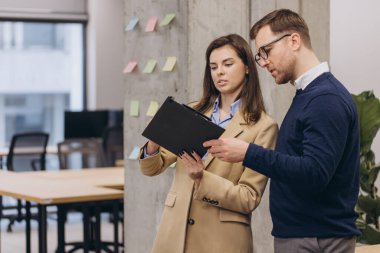 Çeşitli meslektaşlar evrakları inceliyor, yeşil santral ve çağdaş çalışma alanını dekore eden parlak yapışkan notlarla büyük pencerelerin yakınında fikir alışverişinde bulunuyorlar.