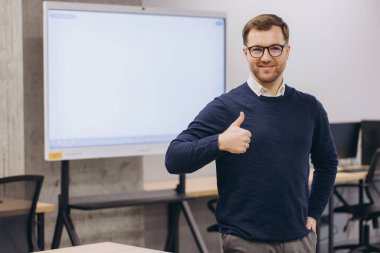 Gülümseyen işadamı onaylama jesti gösteriyor. Modern bir ofis odasında boş bir dijital tahtayla duruyor.