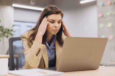 Profesyonel bir kadın şakakları ovuyor, dizüstü bilgisayarla çalışırken gerilimle boğuşuyor. Kurumsal bir çalışma alanında çalışıyor. Yüzünde stres var.