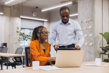 Çağdaş bir ofis ortamında birlikte çalışan çeşitli profesyoneller, bitkiler ve yapışkan notlarla çevrili bir dizüstü bilgisayar ekranına odaklanıyorlar.