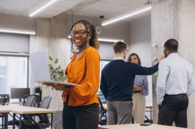 Dizüstü bilgisayarda çalışan Afrikalı Amerikalı profesyonel iş kadını, modern ofis ortamında iş birliği yapan iş arkadaşları, kurumsal takım çalışmasını ve üretkenliği temsil eden