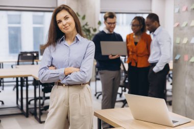 Profesyonel iş kadını kendine güvenerek duruyor, kollarını kavuşturuyor, çağdaş kurumsal çalışma alanında takım olarak birlikte çalışıyor, liderlik ve işbirliğini ön plana çıkarıyor.