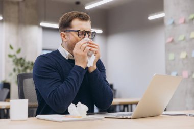 İş adamı gözlük takıyor, ofisteki masasında oturmuş soğuk algınlığıyla boğuşurken burnunu mendile siliyor.