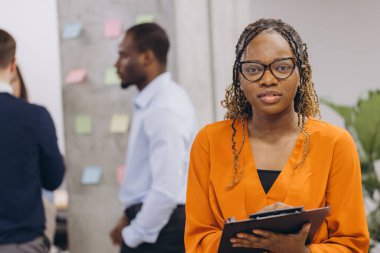 Gülümseyen Afro-Amerikan iş kadını elinde bir klasör tutarken, iş adamları beyin fırtınası yaparken, fikirleri paylaşırken ve modern çalışma alanında birlikte çalışırken, işbirlikçi kurumsal kültürü ön plana çıkarırken,.