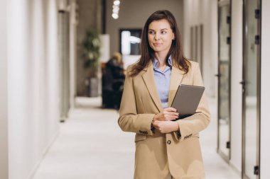 İş kadını güvenli bir şekilde modern ofis koridorunda yürüyor, elinde dijital bir tablet, gülümsüyor ve dikkatlice uzaktan bakıyor.