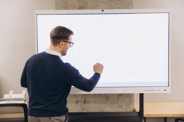 Profesyonel iş adamı interaktif beyaz tahta üzerinde kurumsal verileri analiz ediyor, işbirliği sırasında çağdaş çalışma alanında stratejik grafikler gösteriyor