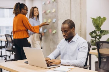 Önplanda dizüstü bilgisayarla çalışmaya konsantre olan Afrikalı Amerikalı iş adamı, arka planda çeşitli ekipler modern ofis ortamını sergileyerek proje üzerinde işbirliği yapıyor.