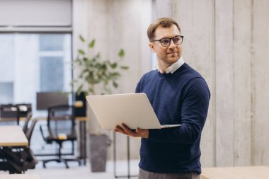 Modern ofis ortamında dizüstü bilgisayar tutan kendine güvenen profesyonel bir adam. Etrafı şık mobilyalar ve büyük pencerelerden gelen doğal ışıklarla çevrili.