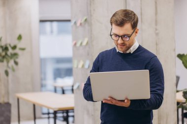 Modern ofis ortamındaki bir adam dizüstü bilgisayarına odaklanmış. Uzaktan kumandalı ya da çevrimiçi çalışmayı işaret ediyor. Çevreleyen elementler işbirlikçi bir ortam öneriyor
