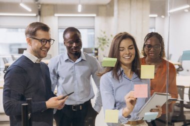 Çeşitli iş adamları, modern ofis ortamında cam bir duvara yapışkan notlar yapıştırarak yaratıcı bir beyin fırtınası yapıyorlar.