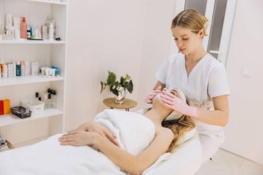 Pembe eldivenli güzellik uzmanı masaj masasında dinlenen sarışın kadına yüz maskesi uyguluyor.