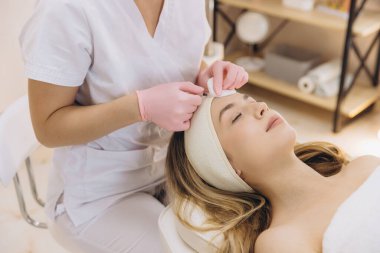 Deneyimli kozmetolog, modern, steril cilt bakımı alanında müşterinin saç çizgisinin yakınına yaşlanma karşıtı yüz enjeksiyonu uyguluyor.