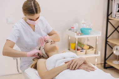 Profesyonel güzellik uzmanı kadının alnına botoks yapıyor, kozmetik yenileme işlemi sırasında koruyucu maske ve eldiven takıyor.