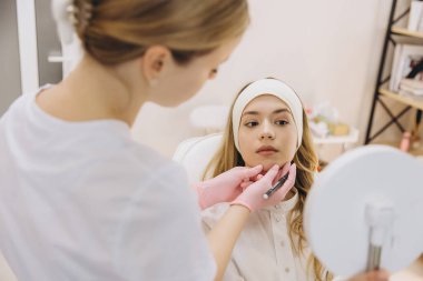 Kozmetik uzmanı genç bir kadının yüzündeki kesin enjeksiyon noktalarını işaretliyor. Profesyonel bir klinikte botoks tedavisine hazırlanıyor.