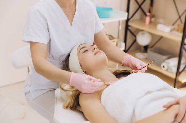 Masaj masasında yatan genç sarışın bir kadının göğsünde fırçayla kozmetik ürünler uygulayan pembe eldivenler giyen bir güzellik uzmanı.