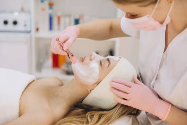 Güzellik uzmanı kaplıcadaki bir müşteriye canlandırıcı yüz maskesi uyguluyor. Ortam temiz ve profesyonel, rahatlama ve cilt bakımını vurguluyor.