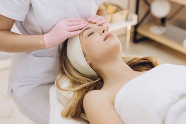 Pembe eldivenli güzellik uzmanı spa ortamında profesyonel cilt bakımı sırasında sarışın müşterinin yüzüne nazikçe masaj yapıyor.