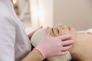 Güzellik uzmanı pembe eldivenler giyiyor. Güzellik salonunda cilt bakımı sırasında genç bir kadının yüzüne krem sürüyor.