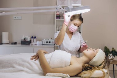 Profesyonel kozmetolog hyaluronik asit enjeksiyonu yapıyor. Sarışın müşteriye yüz gençleştirme tedavisi için lamba ayarlıyor.