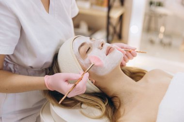 Fırça kullanan bir müşteriye yüz maskesi takan bir spa uzmanı. Rahatlatıcı cilt bakımı kozmetoloji uzmanlığını ve kendine bakımı vurguluyor