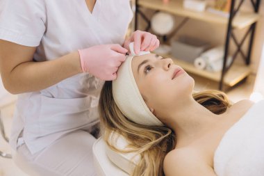 Profesyonel güzellik uzmanı, estetik ameliyattan önce müşterinin alnını dezenfekte ederek modern estetik kliniğinde yüz enjeksiyonuna hazırlanıyor.