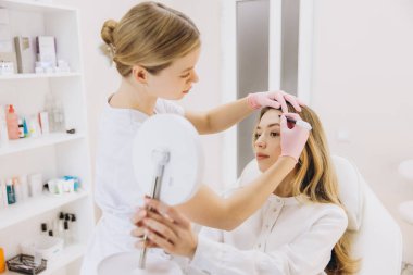 Tıbbi klinikteki kozmetik botoks işlemi sırasında kadın hastanın alnına kesin enjeksiyon bölgelerini işaretleyen profesyonel güzellik uzmanı.