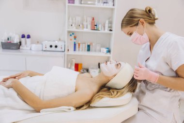 Kaplıcadaki estetisyen bir müşteriye yüz maskesi takıyor. Arka planda cilt ürünleriyle dolu raflar rahatlatıcı bir atmosfer yaratıyor.