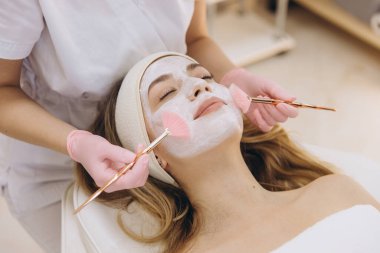 Estetisyen, spa ortamında yüz maskesi takıyor. Müşteri saç bandıyla dinleniyor, cilt bakımı görüyor.