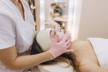 Kozmetik uzmanı, profesyonel bir güzellik merkezinde masaj masasında uzanan genç bir kadının yüzüne canlandırıcı bir maske takıyor.