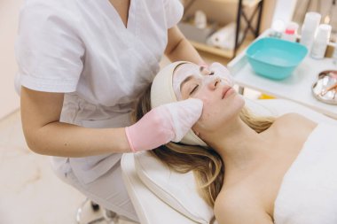 Koruyucu eldiven takan güzellik uzmanı spa tedavisi sırasında rahatlamış bayan müşteriye nazikçe yüz temizleme maskesi uyguluyor.