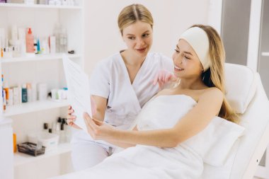 Cilt bakımı belgesini inceleyen profesyonel güzellik uzmanı ve modern spa ortamında rahatlamış müşterisi tedavi yatağında dinleniyor.