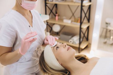 Temiz ve steril bir güzellik salonunda botoks yapmaya hazırlanırken müşterinin alnını dezenfekte eden bir kozmetikçi.
