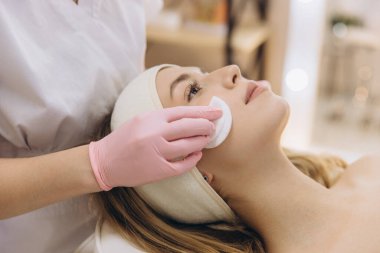 Profesyonel cilt bakımı sırasında bayan müşterinin cildine pamuklu bez ve kozmetik ürünler uygulayan pembe eldivenler giyen bir güzellik uzmanı.