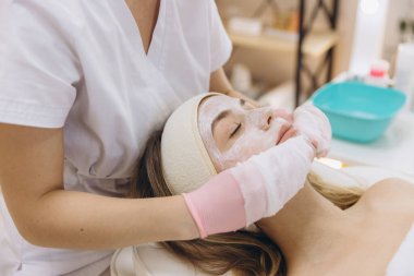 Profesyonel bir güzellik salonunda masaj maskesine uzanmış genç bir kadının yüzüne pembe eldivenler takan bir güzellik uzmanı.