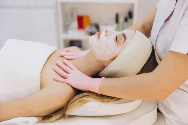 Pembe eldivenli güzellik uzmanı, sakin güzellik stüdyosunda profesyonel spa tedavisi sırasında müşterinin cildine yüz maskesi yayıyor.