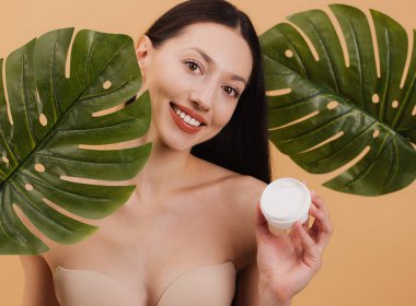 Elinde bir kavanoz nemlendirici krem tutan güzel bir kadın. Arka planda tropikal canavar yaprakları var. Doğal cilt bakımını teşvik ediyor.