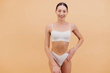 Beyaz iç çamaşırlı, güzel, zayıf Asyalı bir kadının yumuşak bej bir arka plana karşı güvenle poz verdiği bir fotoğraf.