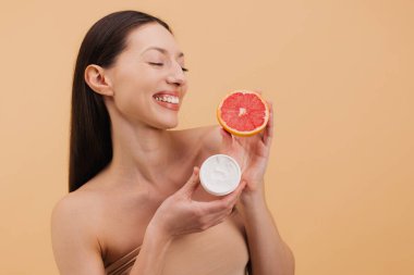 Mutlu kadın, bej arka planda sağlıklı cilt bakımı için greyfurt ve yüz kremi C vitamini karşılaştırıyor.
