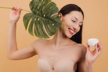 Elinde bir kavanoz nemlendirici ve tropikal yaprak tutan güzel genç bir kadın bej arka planda doğal cilt bakım ürünlerini ve vücut bakımını teşvik ediyor..